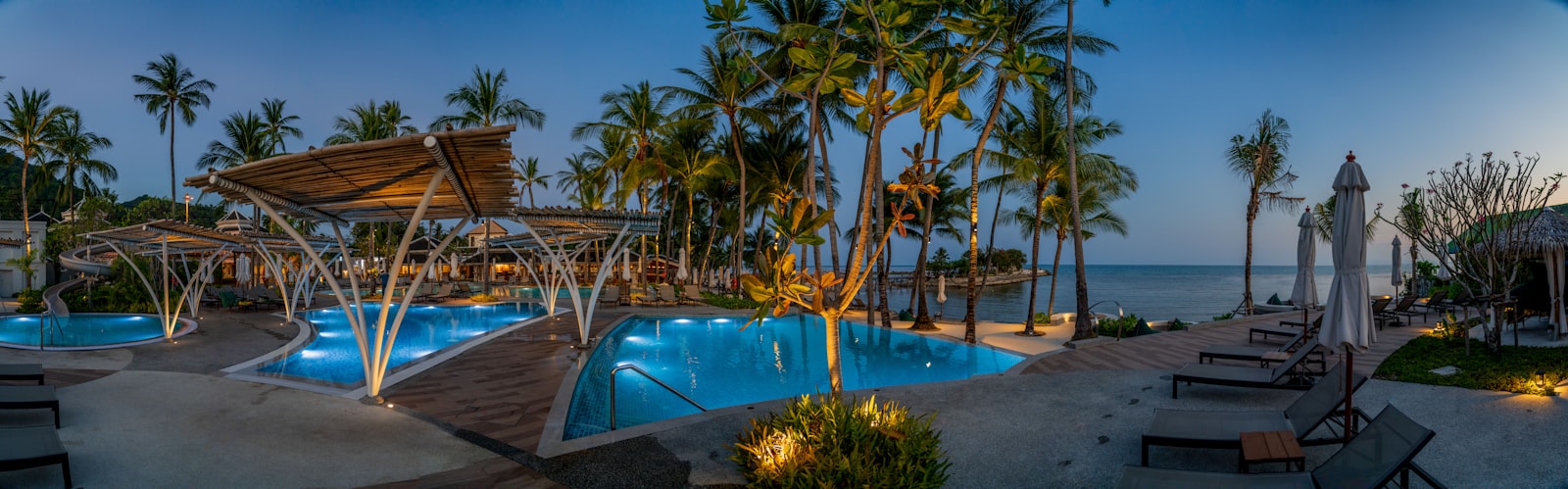 A resort pool deck lit up at dusk with palm trees and ocean views